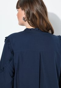 Marineblaue Bluse mit rüschigem Kragen und Ärmelabschlüssen, mit einem Rückenschlitz für einen bequemen Schnitt. Aus leichtem Stoff gefertigt.