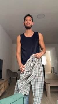 Un jeune homme portant un débardeur noir tient une chemise à carreaux boutonnée et pliée dans un salon moderne.