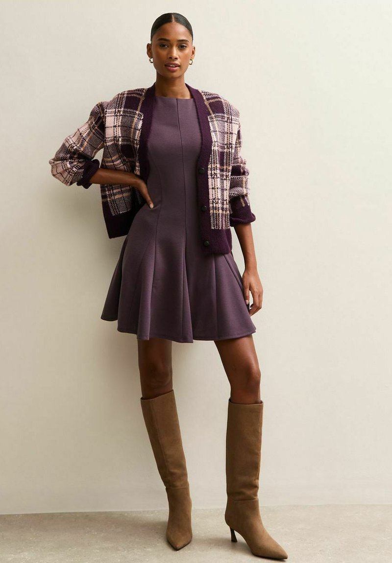 Mujer vestida con un vestido A-line morado, un cárdigan a cuadros en morado y rosa, y botas de tacón de gamuza color marrón claro hasta la rodilla, de pie contra una pared beige lisa.
