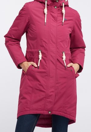 Long manteau rose à capuche avec cordons blancs, poches avant et détails de boutons porté sur un pantalon bleu foncé.