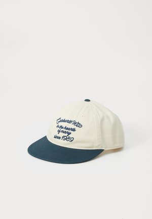 Casquette avec une couronne en coton crème et un bord bleu marine ; comporte un texte brodé "Carhartt WIP" et l'année "1989". Texture douce.