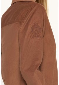 Veste en velours côtelé marron avec col, présentant un emblème brodé sur la manche gauche, une texture subtile et une coupe décontractée.