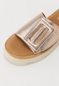 Gros plan d'une sandale slide en or rose métallique avec une large bride et une boucle rectangulaire surdimensionnée sur une semelle beige rembourrée.
