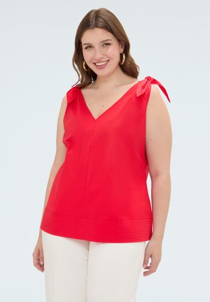Donna che indossa un top rosso senza maniche con spalline annodate e pantaloni bianchi, sorridente davanti a uno sfondo chiaro e uniforme.