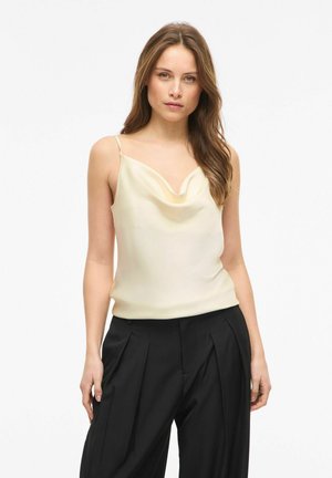 Frau mit langen braunen Haaren, die eine cremefarbene Seiden-Cowl-Neck-Camisole und hoch taillierte schwarze plissierte Hosen trägt, stehend vor weißem Hintergrund.