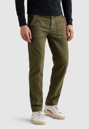 Man draagt olijfgroene broek met voorzakken en witte sneakers, staand tegen een effen lichte achtergrond.