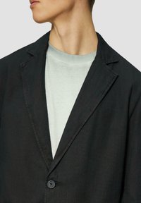 Schwarzer Blazer aus strukturiertem Stoff mit Reverskragen und Knopfverschluss; über einem hellgrauen Rundhals-Shirt getragen.