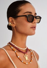 Γυναίκα με τα μαλλιά τραβηγμένα πίσω, που φοράει μαύρα γυαλιά ηλίου, πολλαπλά χρυσά σκουλαρίκια που αγκαλιάζουν το αυτί και πολλά ενωμένα κολιέ με χάντρες, μεταξύ των οποίων ένα με χρυσό μενταγιόν σε σχήμα χελώνας.