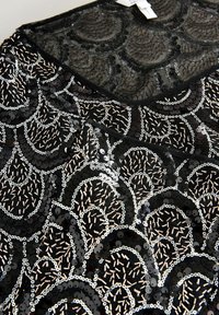 Čierny top s flitrami, ktorý má textúrovaný dizajn s kruhovými vzormi, kovovými akcentmi a priehľadným výstrihom z ľahkej látky.