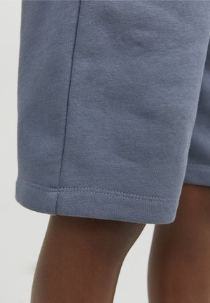 Persona de piel oscura vistiendo pantalones cortos de punto azules de longitud media, mostrando la costura del dobladillo y la textura de la tela contra un fondo claro y liso.