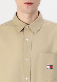 Chemise beige à manches longues en coton, avec un col classique, des boutons blancs et une poche sur la poitrine gauche arborant le logo "Tommy Jeans".