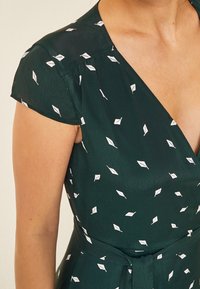 Robe vert foncé à manches courtes, design croisé et motifs de feuilles blanches ; elle dispose d'une ceinture à nouer à la taille et d'une texture lisse.