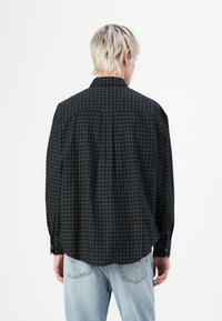 Chemise à carreaux noirs à manches longues, avec un motif de grille blanche et un détail de pli dans le dos. Fabriquée en tissu léger, avec des boutons visibles aux poignets.