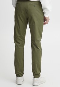 Olivenfargede slim-fit bukser laget av lett materiale med to baklommer og en enkel knappelukking. Hvite joggesko er på føttene.