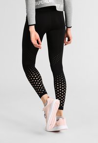 Osoba v černých perforovaných legínách s vzorovanými otvory na lýtkách a bledě růžových teniskách, stojící na šedém povrchu.