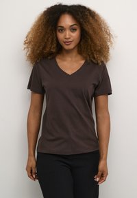 T-shirt a V col marrone realizzato in tessuto morbido. Maniche corte, vestibilità rilassata e texture liscia messa in evidenza su uno sfondo neutro.