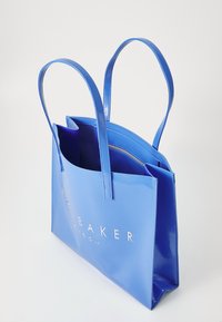 Modrá lakovaná kabelka s dvoma uchami, ktorá má priestranný interiér a embosované logo na prednej strane. Hladký povrch, vrátane vertikálnych vreciek.