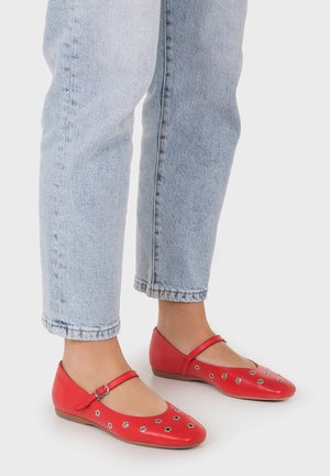 Pies con vaqueros azul claro y zapatos planos Mary Jane rojos con pequeños patrones florales recortados sobre fondo blanco.