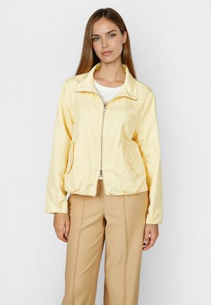 Woman wearing a light yellow zip-up jacket over a white shirt, paired with beige pants, standing against a plain white background.