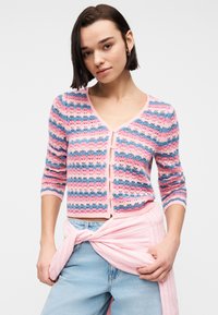 Γυναίκα που φοράει ριγέ πλεκτή ζακέτα σε ροζ, μπλε και εκρού, ανοιχτό μπλε τζιν και ροζ πουκάμισο δεμένο στη μέση της, σε λευκό φόντο.