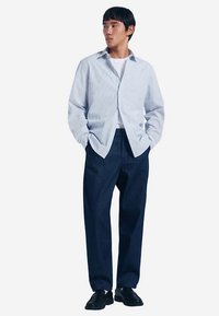 Vaaleansininen raidallinen nappipaita, yhdistetty laivastonsinisiin leveälahkeisiin housuihin. Asun täydentävät mustat kengät. Malli seisoo käsineen taskuissa.