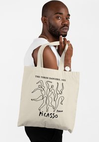 Beżowa torba na ramię z płótna z czarnym rysunkiem przedstawiającym tańczące postacie. Napis brzmi "TRZY TANCERKI, 1925" oraz "PICASSO."