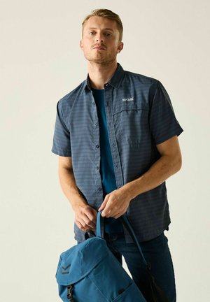 Mann trägt ein blau gemustertes Kurzarmhemd und dunkle Jeans und hält einen blauen Rucksack an den Trägern vor hellem Hintergrund.