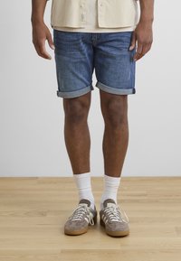Shorts en denim avec un lavage bleu et des ourlets retroussés, portés avec des chaussettes blanches et des baskets en daim marron avec des rayures blanches.