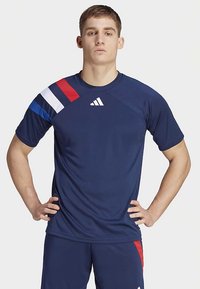 Tengerészkék sportpóló rövid ujjakkal, egyik vállán piros és fehér átlós csíkokkal, valamint fehér Adidas logóval.