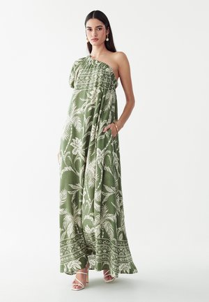 Mujer con un vestido maxi verde de un solo hombro con patrones florales blancos y sandalias blancas de tacón, de pie frente a un fondo liso.