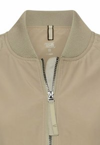 Beżowa kurtka typu bomber z prążkowanym kołnierzem, centralnym srebrnym zamkiem błyskawicznym oraz wewnętrzną metką z marką "camel active", rozmiarem 38 i napisem "recycled".