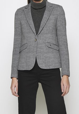 Kvinde iført en grå ternet blazer over en mørk rullekrave og sorte bukser, stående mod en ensfarvet lys baggrund.