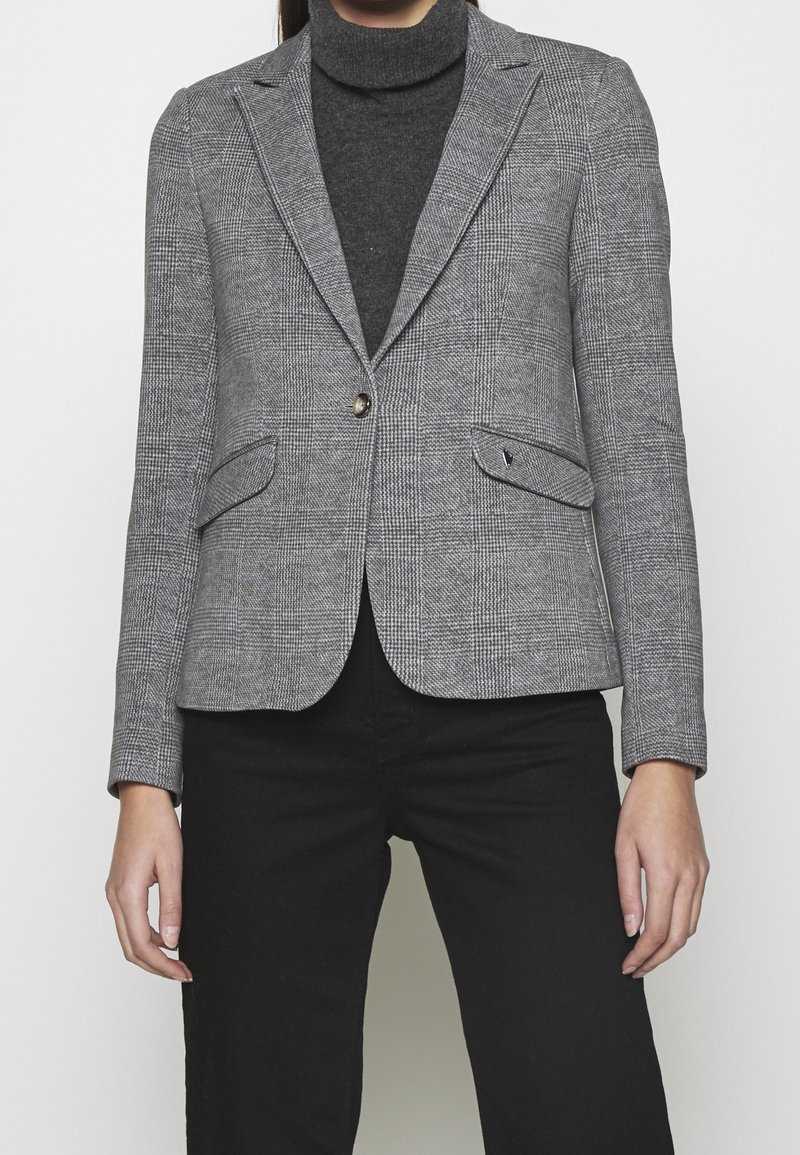 Femme portant un blazer gris à carreaux par-dessus un pull à col roulé foncé et un pantalon noir, debout devant un fond clair uni.