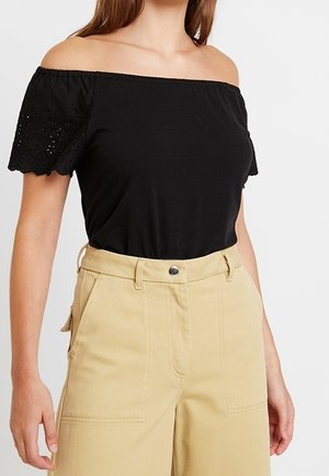 Vrouw draagt een zwarte off-shoulder top met kanten mouwen en beige wijde broek met hoge taille tegen een witte achtergrond.