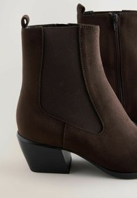 Brown suede ankle boots with elastic side panels, distressed texture, and a stacked black heel. Features a zipper for easy wear.