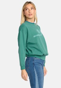 Groene katoenen sweatshirt met een ronde halslijn en lange mouwen, voorzien van een witte grafische print. Gecombineerd met blauwe denim jeans.