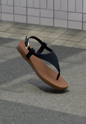 Sort læder tåsandale med brun sål og guldbælte svæver over asfalt ved siden af flisebelagt væg.