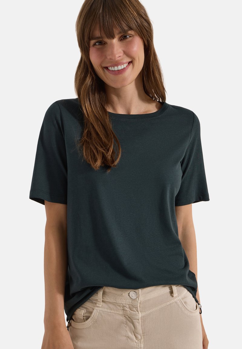 Femme souriante portant une chemise à manches courtes vert foncé et un pantalon beige, les mains dans les poches devant un fond uni.