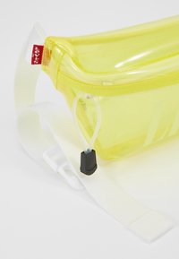 Sac banane transparent jaune avec sangle blanche réglable, tirette de fermeture éclair noire et étiquette rouge de la marque Levi’s sur un fond clair.