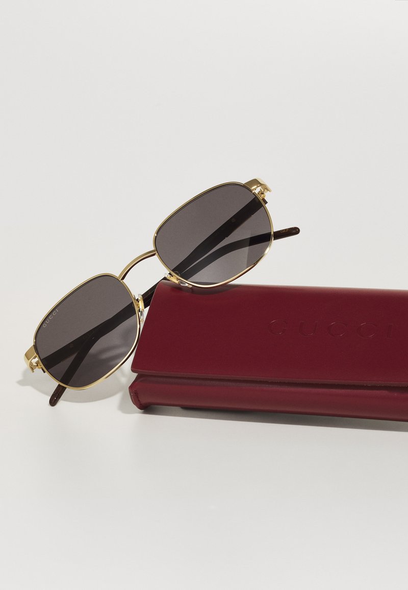 Lunettes de soleil à monture dorée avec des verres sombres, présentées sur un étui en cuir rouge profond embossé avec "GUCCI." Forme de lentille carrée et design élégant.