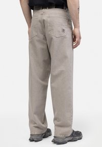 Pantalones de mezclilla beige con un ajuste relajado, que cuentan con dos bolsillos traseros, costuras visibles y una etiqueta. Llevados con zapatillas grises.