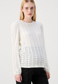 White long-sleeve blouse with puffed shoulders, featuring a textured design of vertical stripes and small cutouts throughout.