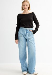 Suéter negro de punto con hombros al descubierto y pantalones cargo de mezclilla azul claro de pierna ancha con bolsillos laterales. La modelo se encuentra contra un fondo blanco liso.