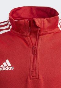 Sudadera roja con cremallera y cuello alto, con franjas blancas en los hombros. Presenta un pequeño bolsillo con cremallera y un logotipo bordado.