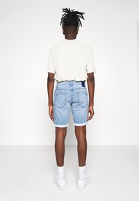 Pantalones cortos de mezclilla azul claro con dobladillos enrollados, etiqueta azul en la cintura y dos bolsillos traseros. Combinados con una camiseta color crema y zapatillas deportivas blancas.