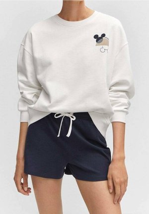 Osoba v bílém svetru s malým motivem Mickey Mouse a tmavě modrých šortkách s šňůrkou v pase, stojící s uvolněnýma rukama.