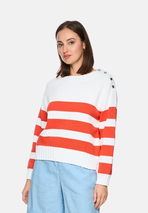 Woman with shoulder-length brown hair wears white sweater with wide orange stripes and light blue pants, standing against plain background.