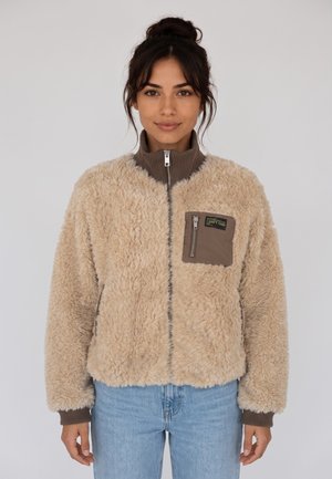 Giovane donna con capelli scuri raccolti in uno chignon, indossa una giacca beige soffice con cerniera, con una tasca sul petto marrone con cerniera, e jeans blu.