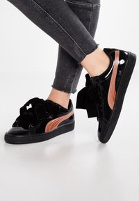 Nohy v černých skinny džínech a lesklých černých teniskách se růžově zlatými pruhy a velkými černými mašlemi na bílém podlaze.