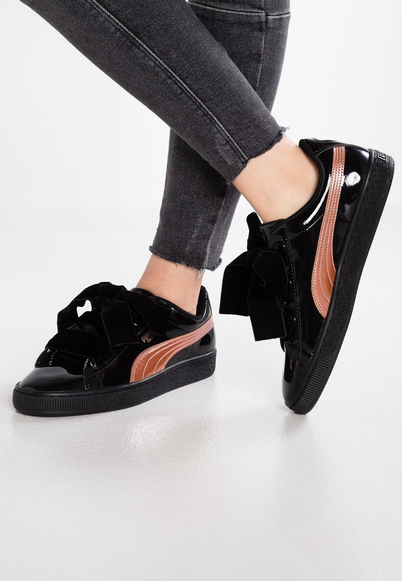 Nohy v černých skinny džínech a lesklých černých teniskách se růžově zlatými pruhy a velkými černými mašlemi na bílém podlaze.